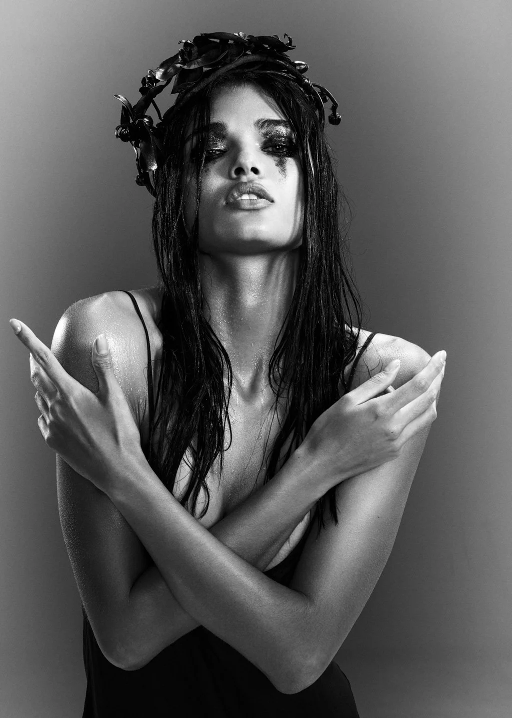 Black and white portrait of a woman with wet hair, wearing a headpiece made of ribbons, crossing her arms over her chest, with water droplets on her skin, and a confident expression.