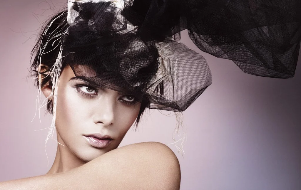 A woman with short hair wearing a large black tulle headpiece against a pink background.