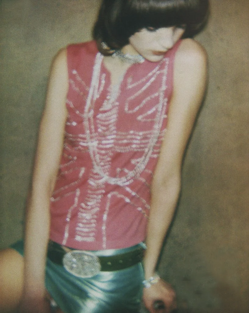 A woman with dark hair wearing a sleeveless red top with a white pattern, a large belt buckle, and teal shorts.