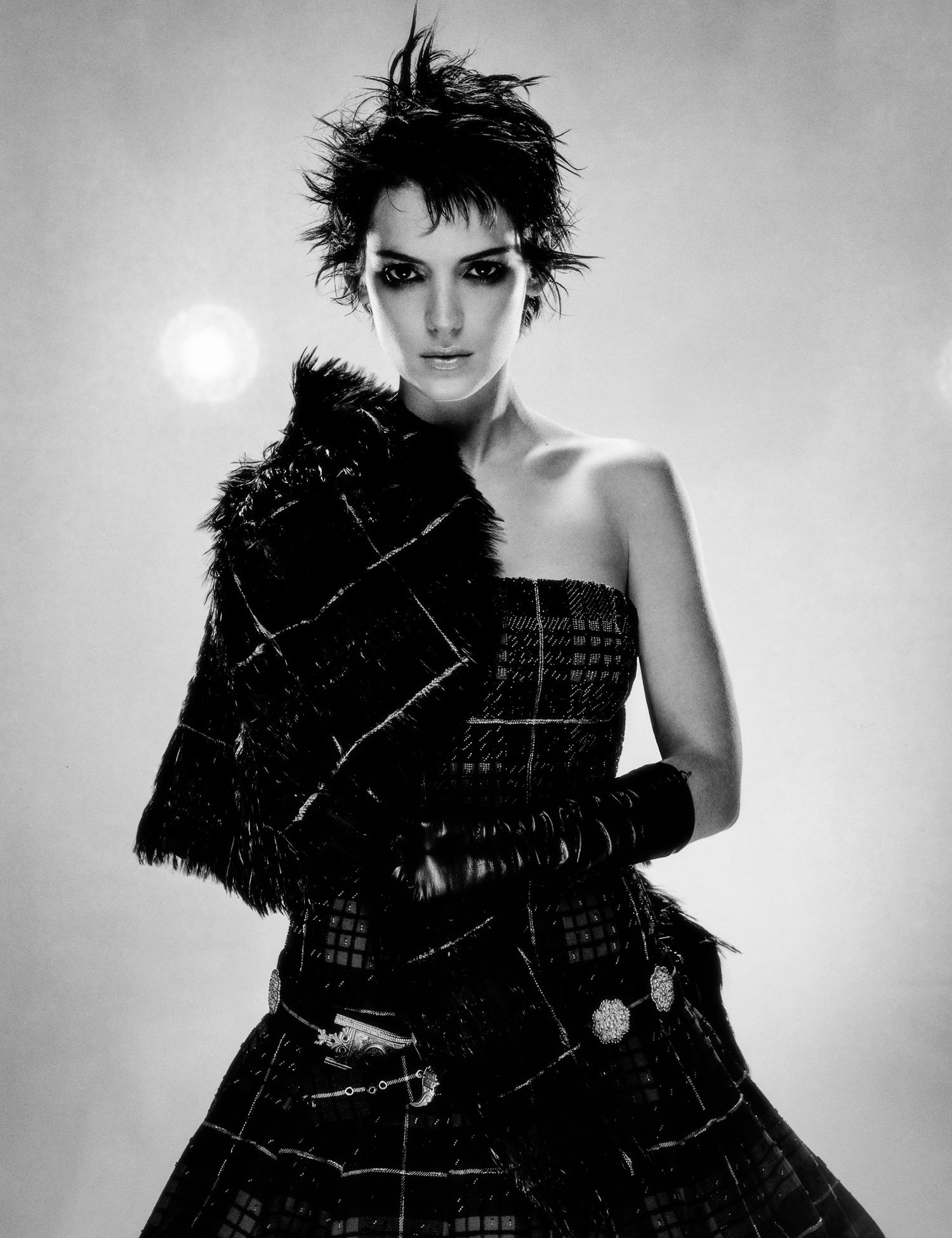 A black and white photo of WINONA RYDER with spiky short hair, wearing a patterned strapless dress, a fur jacket draped over her shoulder, and long gloves, looking directly at the camera with a serious expression.