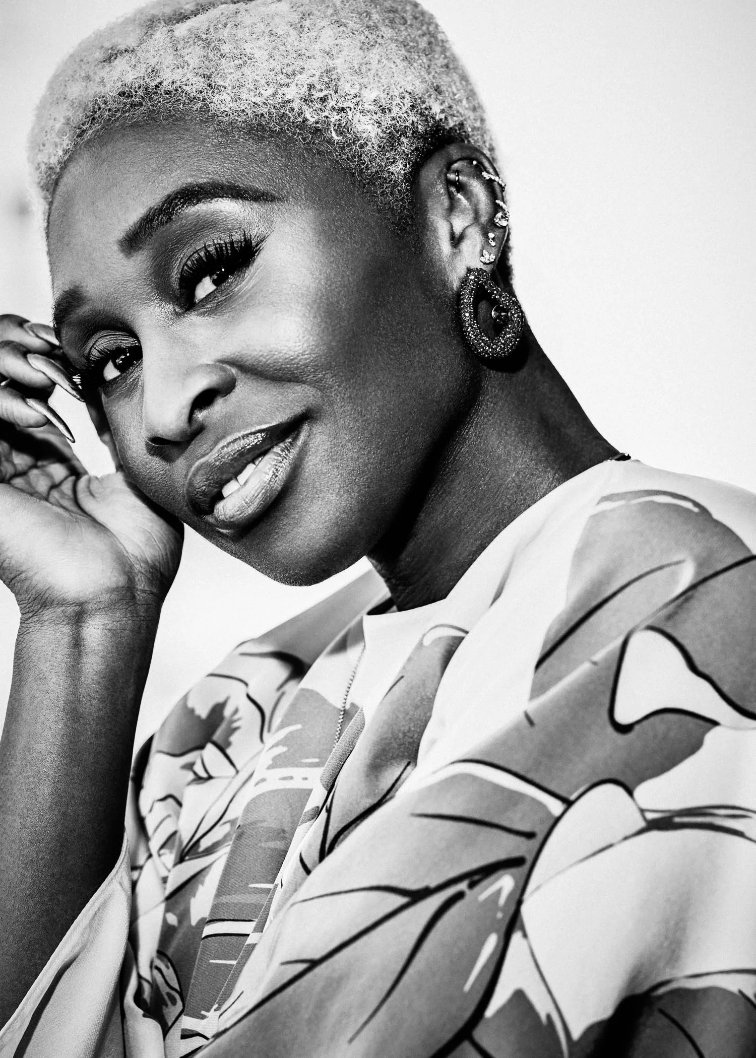 A black and white portrait of a woman with short, platinum blonde hair, wearing earrings and a patterned top, posing with her head tilted and her hand near her face.