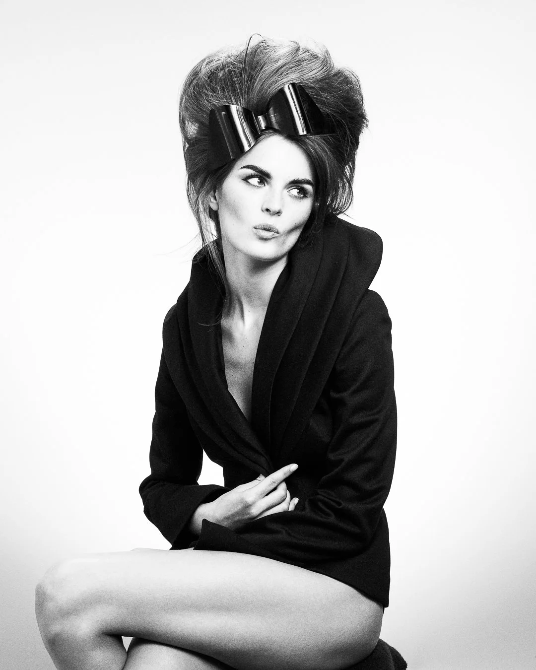 Black and white studio portrait of a woman with voluminous hair styled with large hair rollers, wearing a black blazer with the collar up, sitting with one leg crossed over the other, looking thoughtfully to the side.