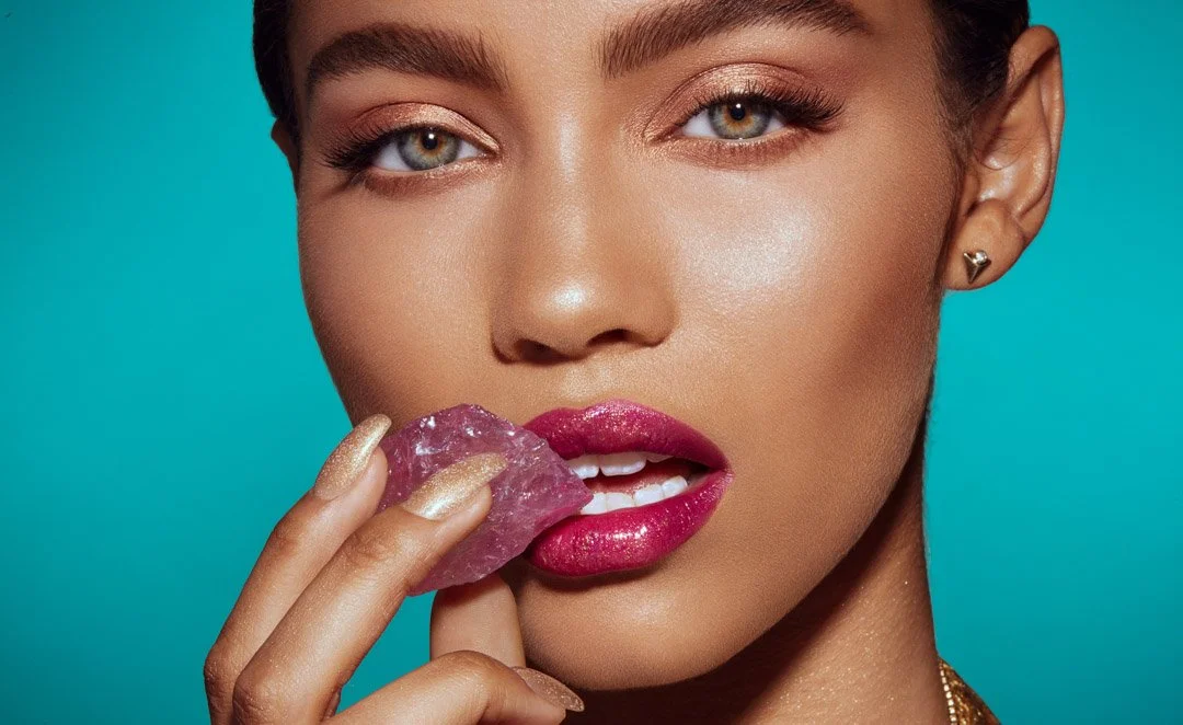 A woman with bright makeup and gold nail polish is holding a pink crystal near her lips.