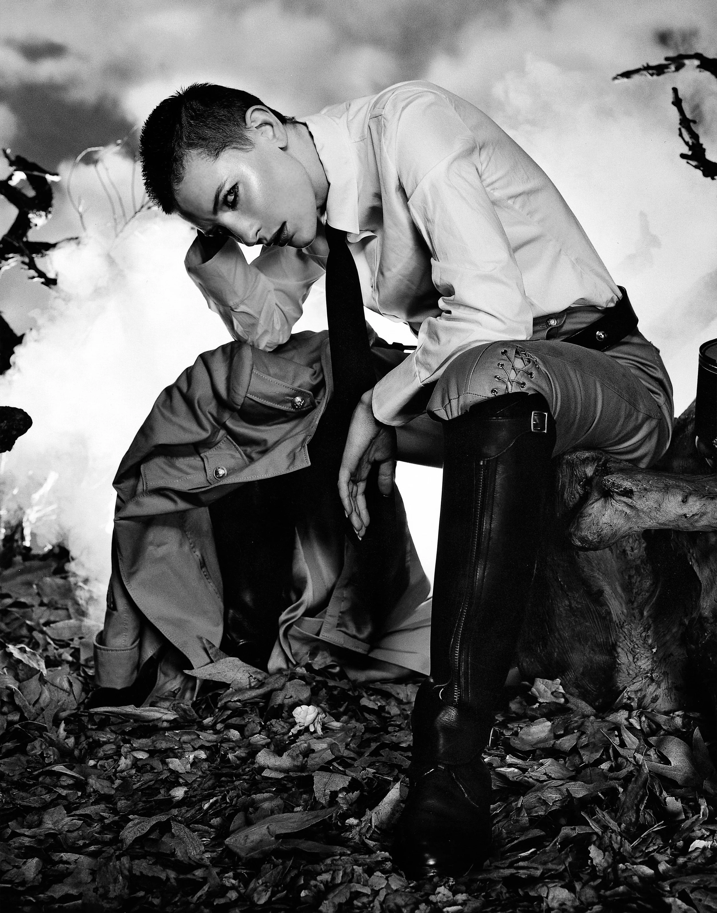 Black and white photo of CATE BLANCHETT with short dark hair, wearing a shirt, tie, and tall boots, kneeling on a bed of leaves outdoors, with a cloudy sky in the background.