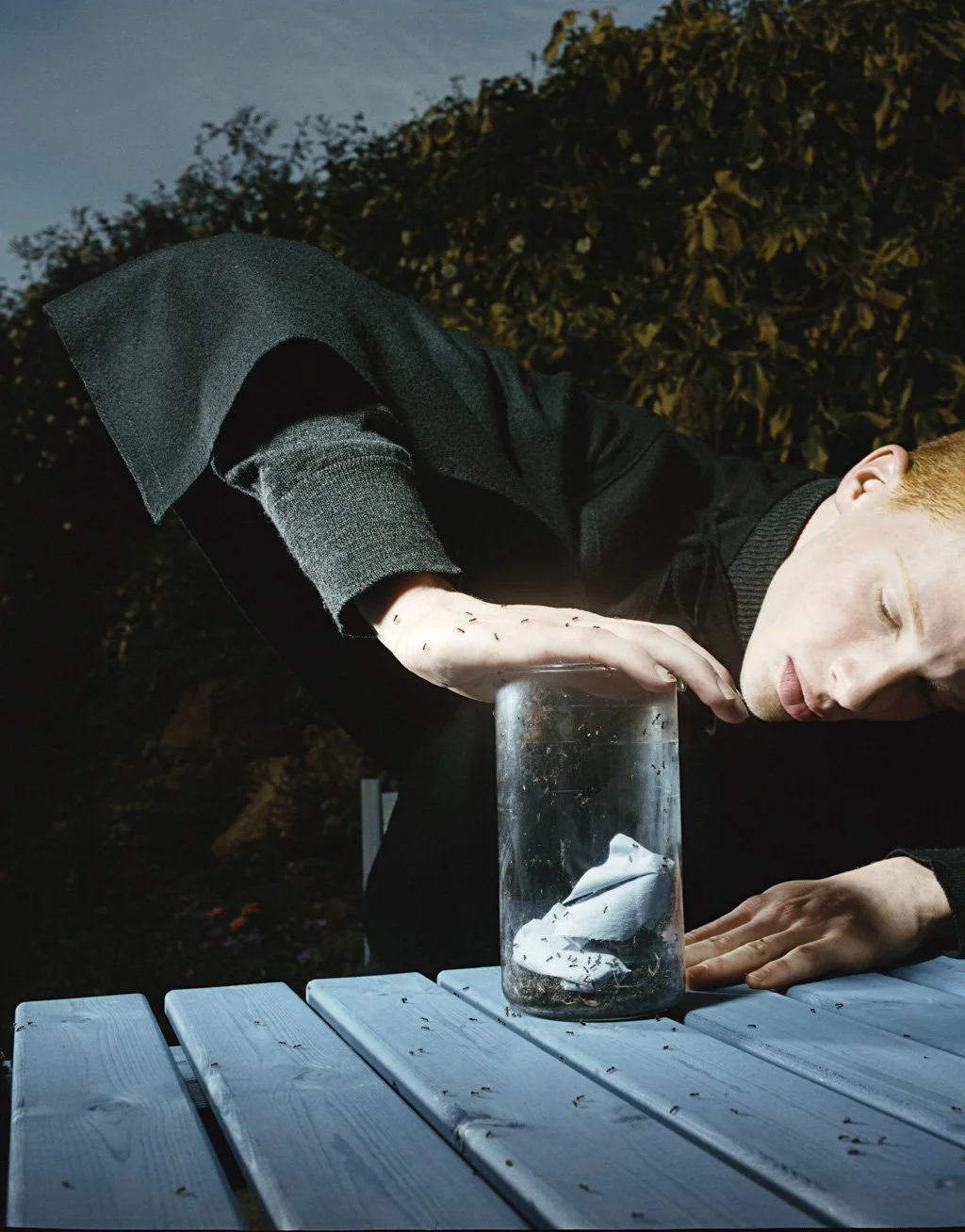 A person with red hair and pale skin is leaning over a glass jar on a blue wooden table outdoors at dusk, with trees in the background. The person appears to be smelling the jar, which contains crumpled paper and dirt. The person is wearing a black j