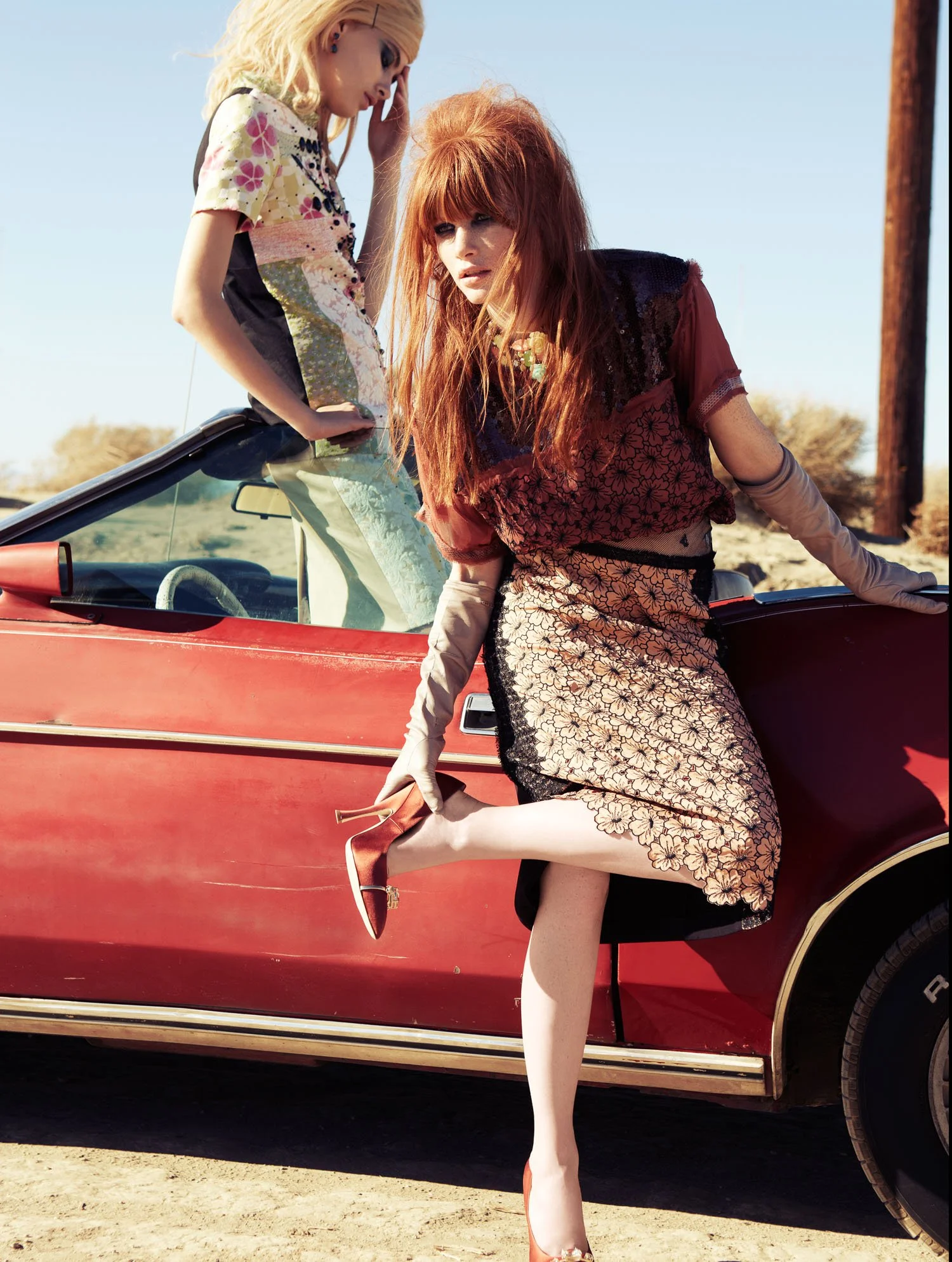 Two women posing by a vintage red convertible car outdoors with a clear blue sky and desert landscape in the background. One woman with red hair and floral dress leaning on the car's door, the other woman with blonde hair and colorful top standing on