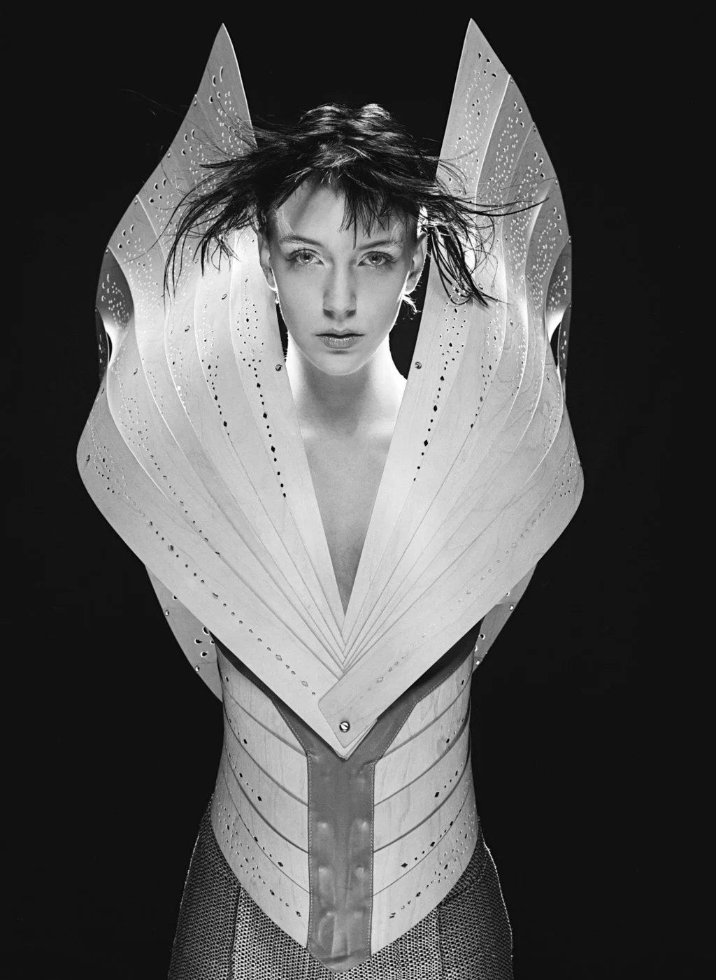 A black-and-white photograph of a woman with short, tousled hair and a serious expression, wearing an avant-garde sculptural costume with large, leaf-like structures around her shoulders and a fitted bodice with textured patterns.