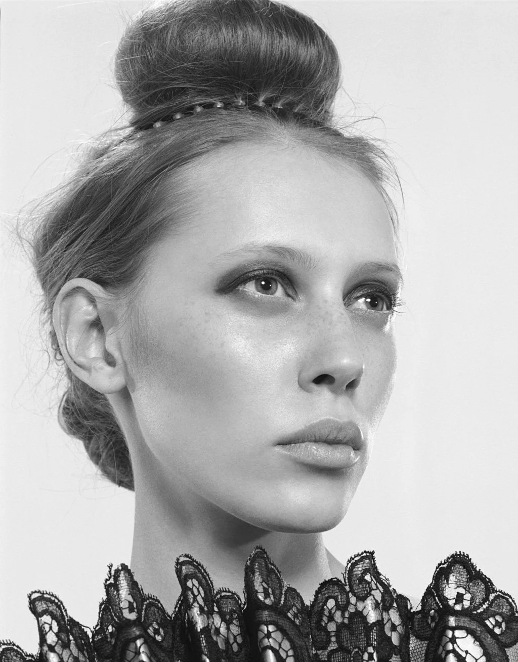 Black and white close-up portrait of a woman with styled hair in a bun, wearing a lace top, looking slightly upward.