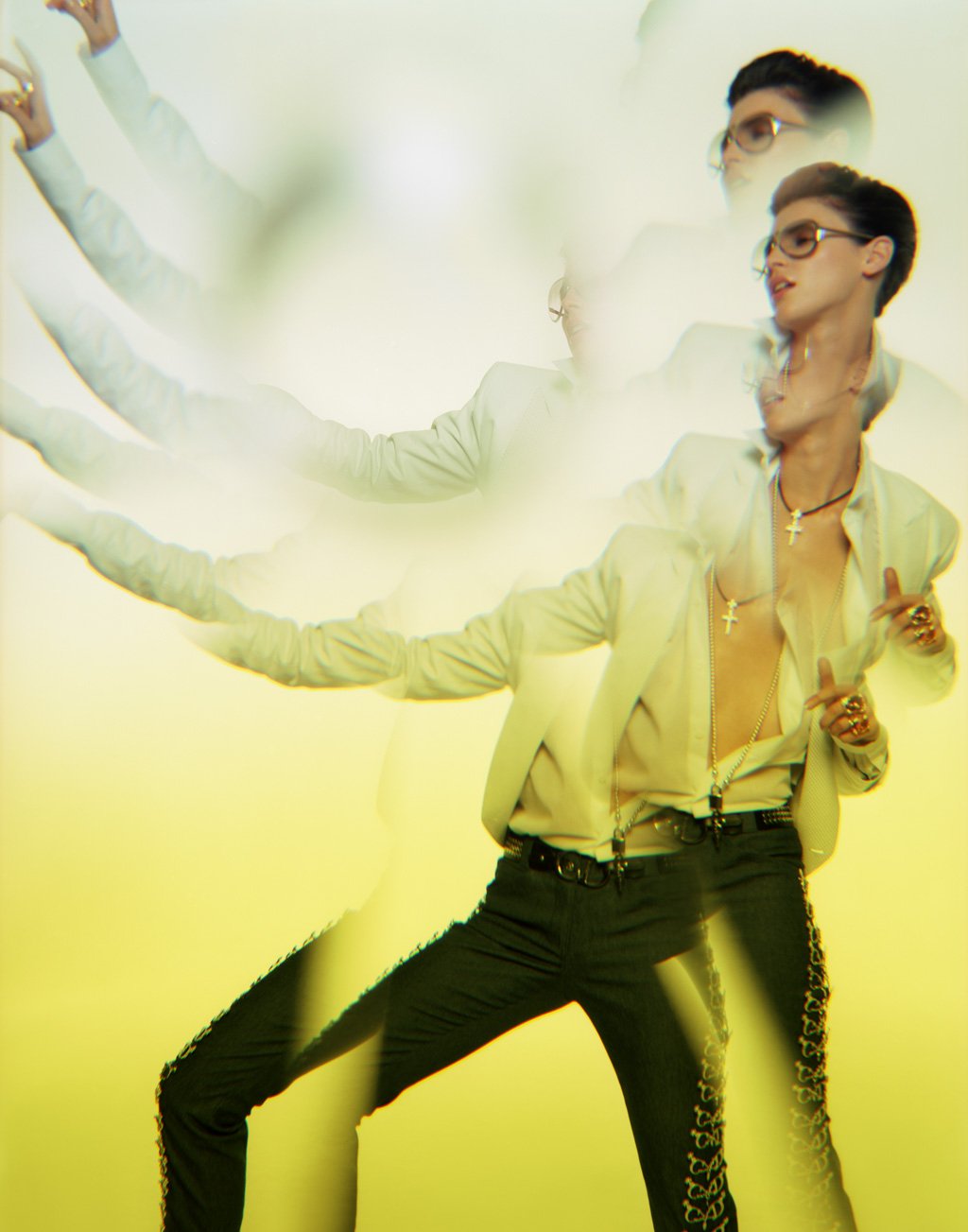 A photo with multiple exposures of a woman with short dark hair, wearing glasses, a yellow blazer, open shirt, multiple necklaces including crosses, and black pants with gold embroidery, holding a cigarette, against a yellow background.