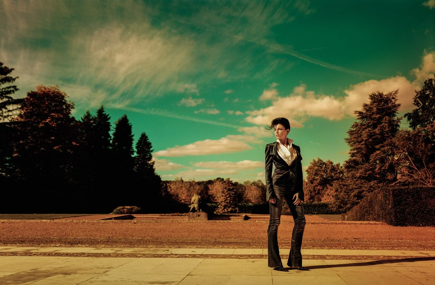 A woman with short dark hair wearing a black suit with a white shirt standing outdoors on a paved surface with a park and trees in the background under a greenish sky with clouds.