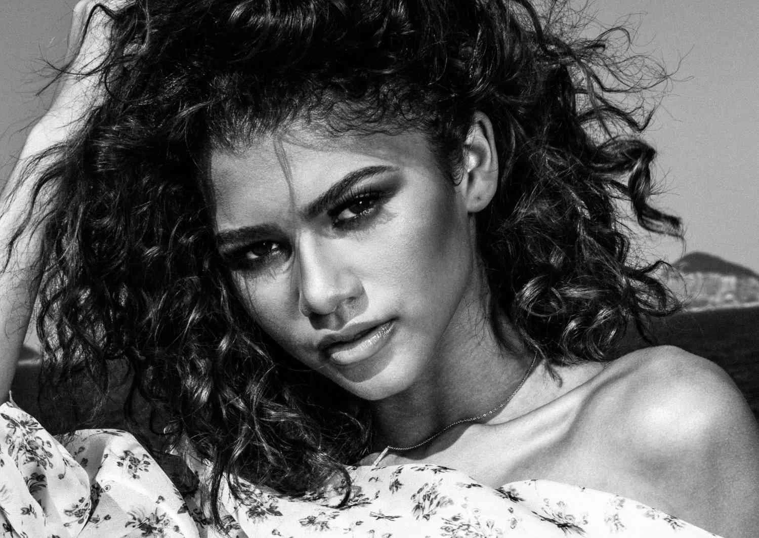 A black and white close-up portrait of ZENDAYA with curly hair, partially covered shoulder, intense gaze, and a floral fabric visible on her arm.