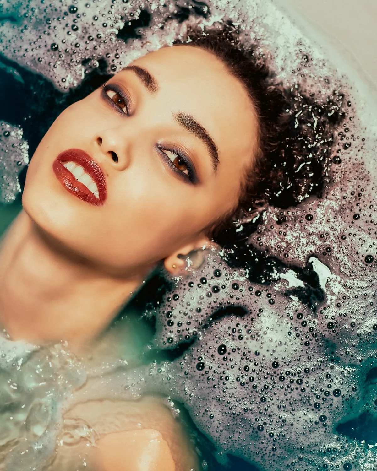 Close-up of a woman's face with makeup, lying in a bubble-filled bath with foam and bubbles surrounding her head.