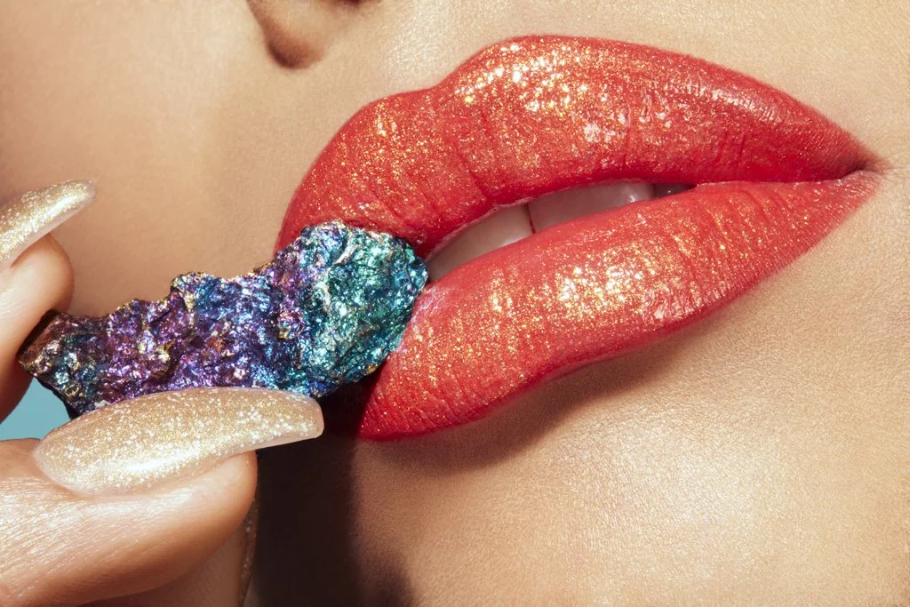 Close-up of a woman's lips painted with shimmery red lipstick, with a hand holding a colorful, textured rock near her lips, and gold-glittered fingernails.