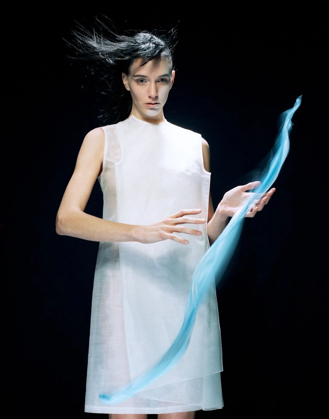 A woman with black hair and light skin wearing a white sleeveless dress, holding a glowing blue ribbon, against a black background.