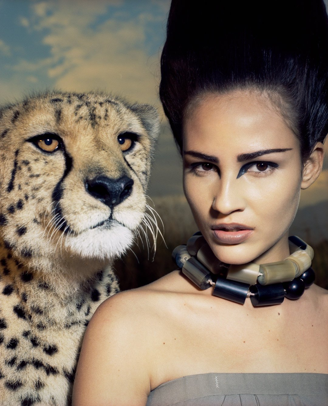 A woman with styled hair and bold makeup, wearing a chunky black and beige necklace, stands next to a cheetah with a spotted coat, both looking into the distance with a landscape background.