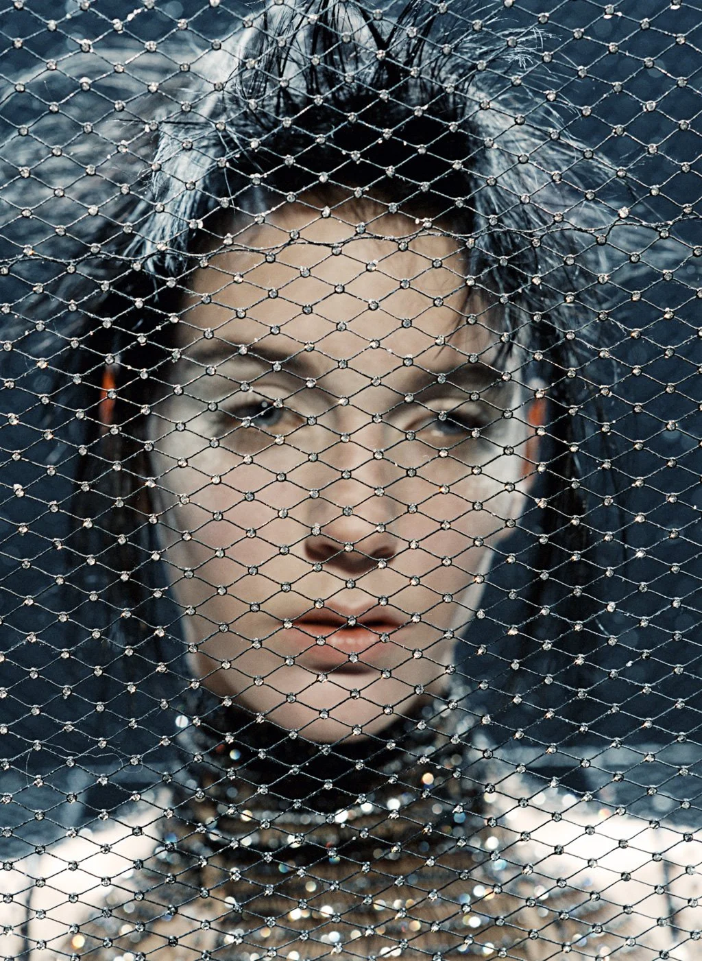 A woman with dark hair and makeup behind a diamond-patterned chain-link fence.