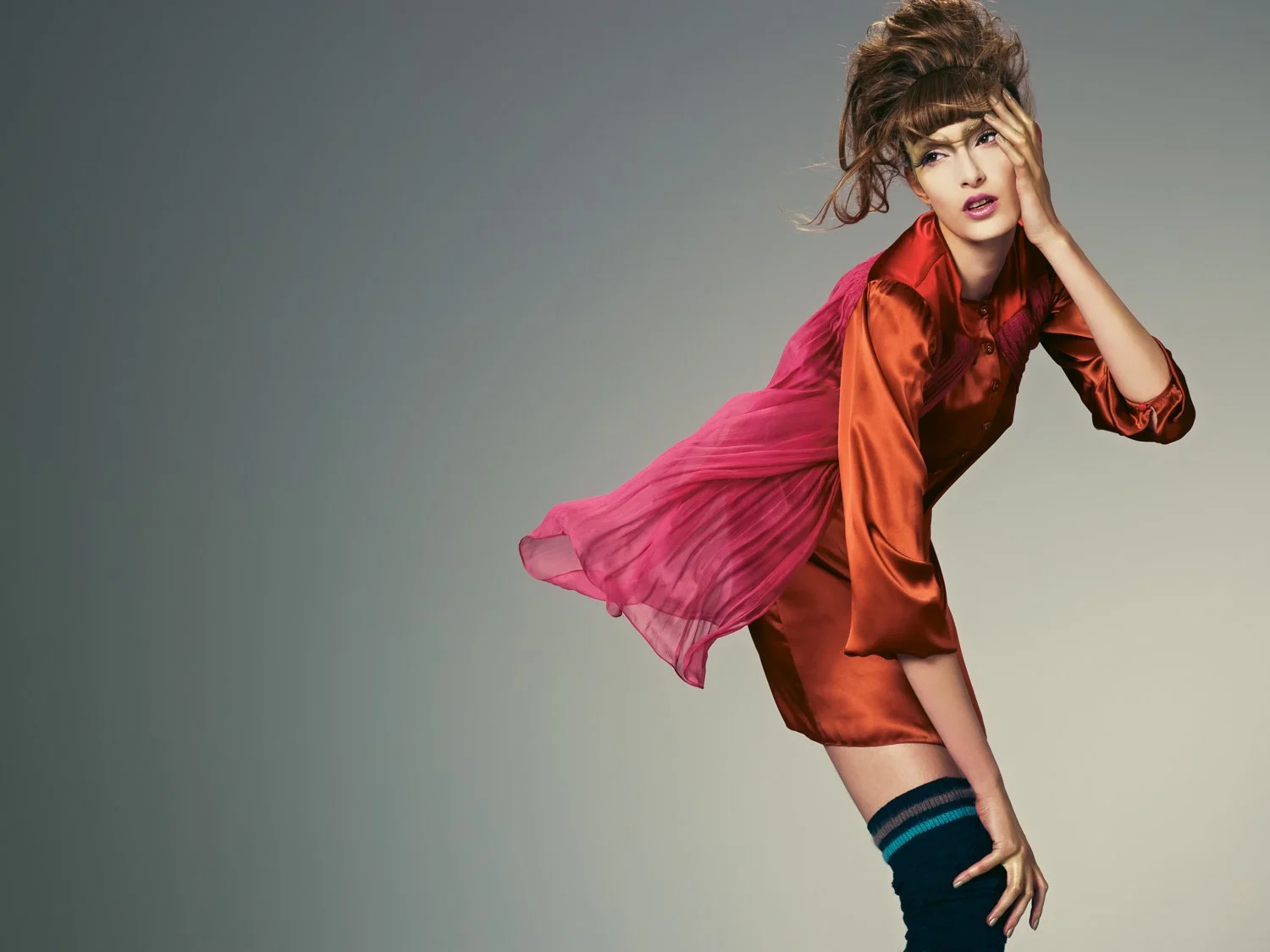 A woman with curly brown hair wearing a shiny orange satin dress with a pink sheer overlay, black knee-high socks, and striking makeup poses against a plain, gradient background.