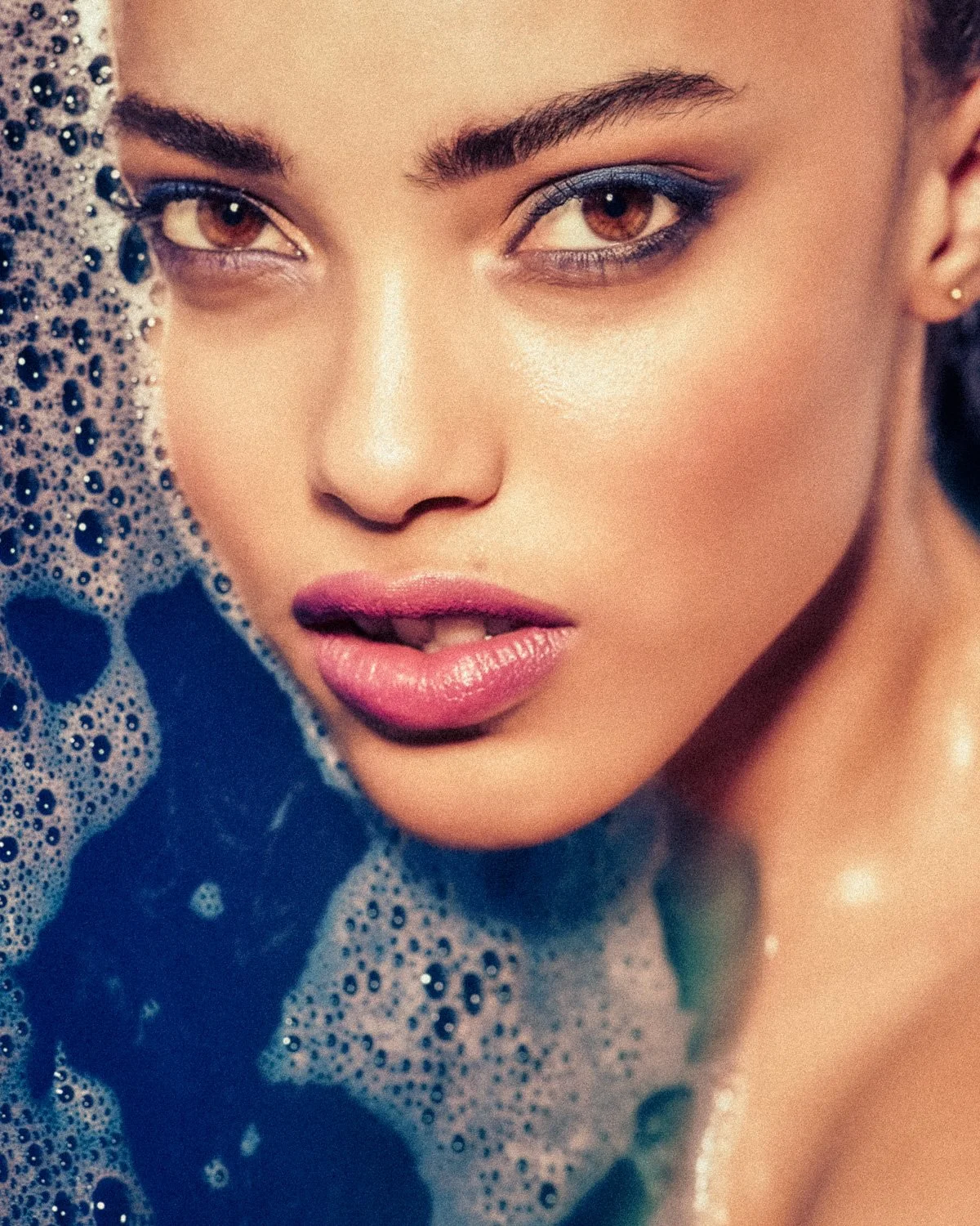 Close-up of a woman with brown eyes and makeup, partially submerged in soapy water with bubbles.