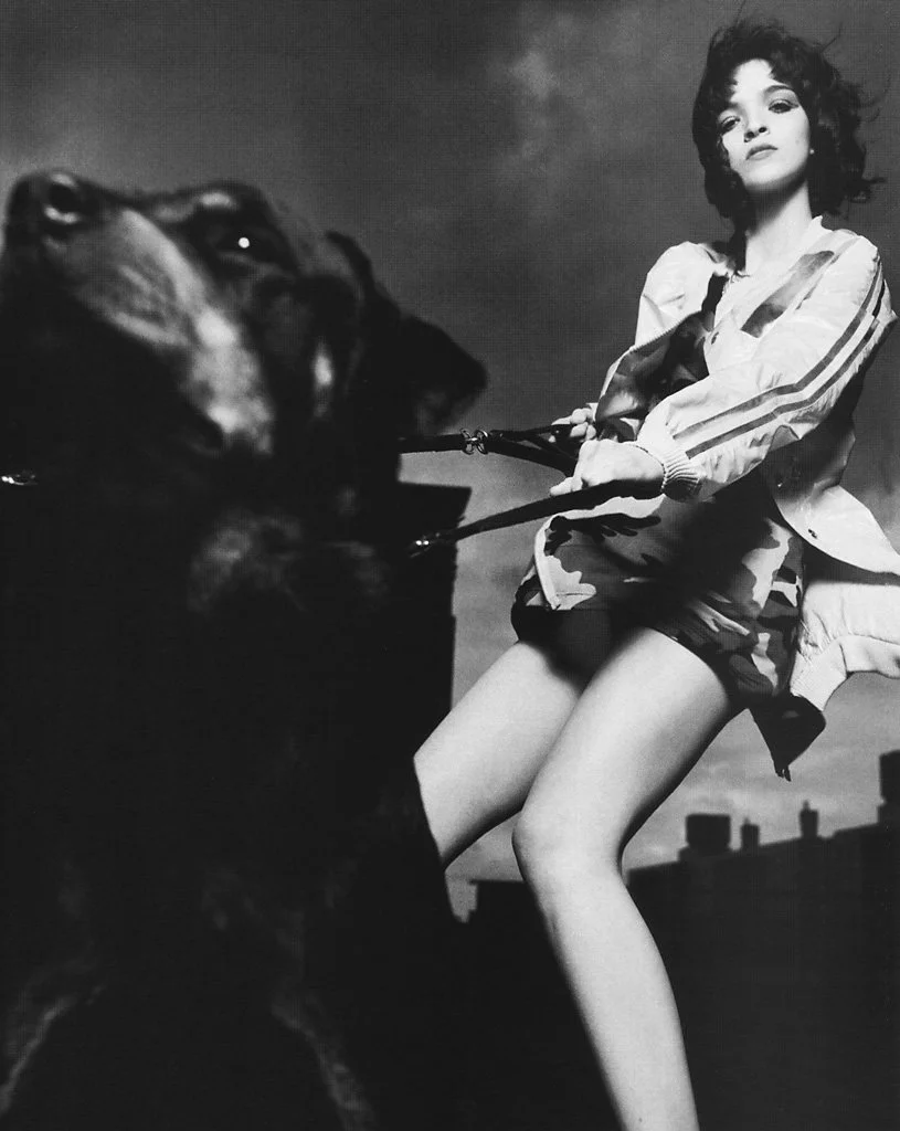 A woman in a casual jacket and shorts outdoors, holding a leash of a large dog, with rooftops in the background and cloudy sky.