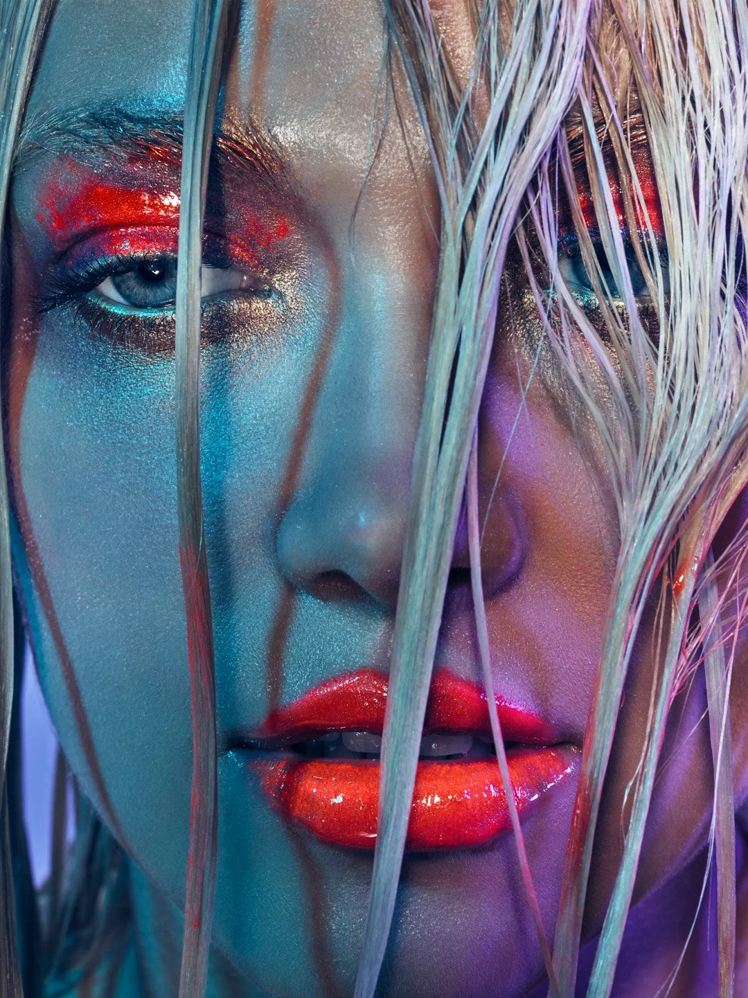 Close-up of a woman's face with wet hair, vibrant makeup with red eyeshadow and glossy red lipstick, surrounded by colorful lighting.