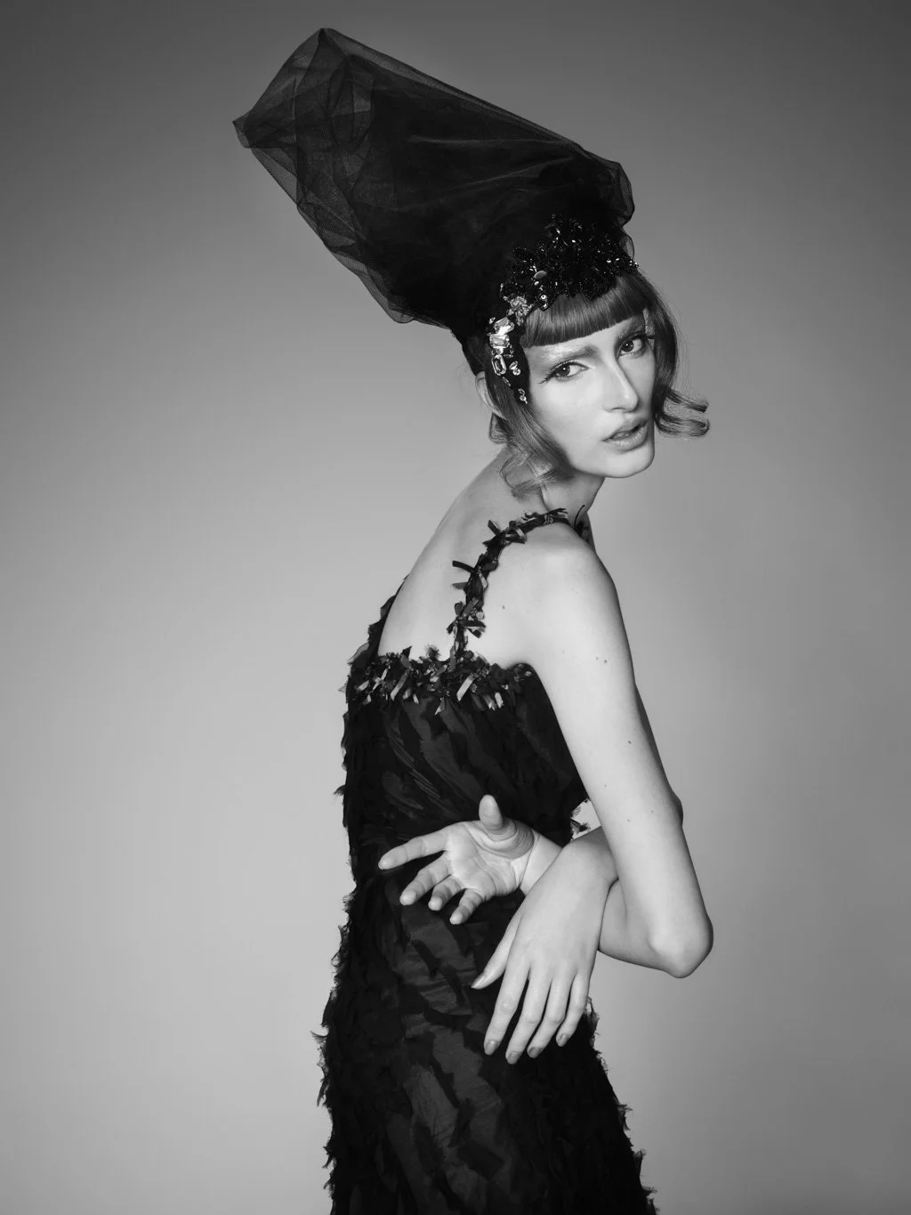 A black and white photo of a woman wearing an elaborate headpiece and a dress with decorative shoulder straps, with her arms crossed and her head slightly tilted.