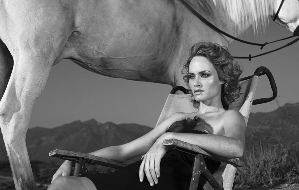 A woman with wavy hair sitting in a reclining chair outdoors, with a horse standing beside her. Mountains are visible in the background.