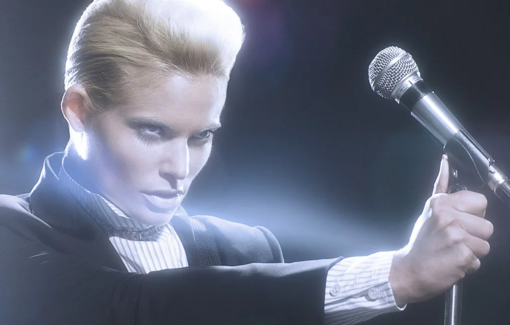 A person with slicked-back blonde hair holding a microphone on a stand, wearing a black blazer and a white shirt with stripes, looking intensely at the microphone.