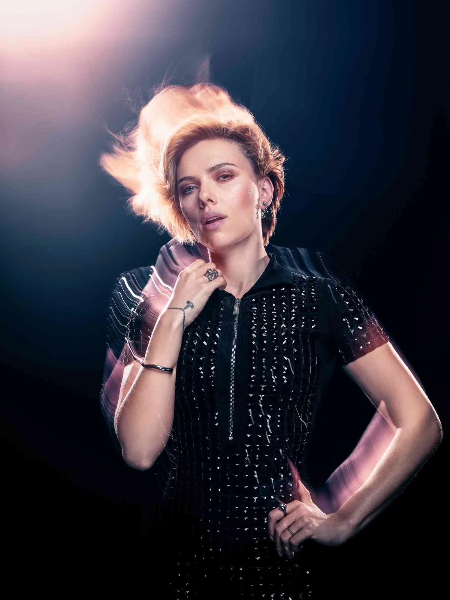 SCARLETT JOHANSSON with short, styled hair and multiple earrings poses in front of a dark background with pink and purple light effects.