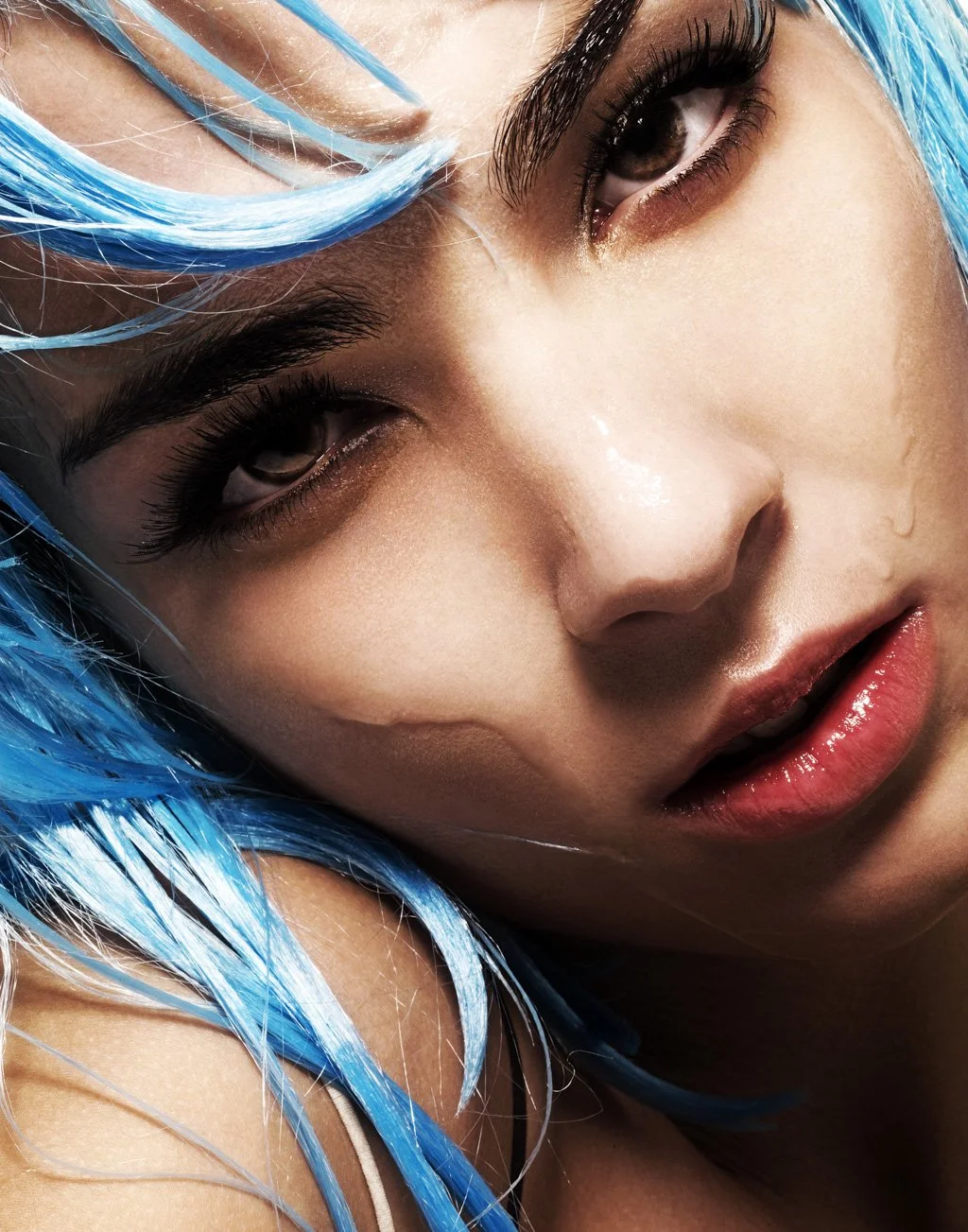 Close-up of a woman with vibrant blue hair, dark eye makeup, and glossy lips, leaning her face on her arm.
