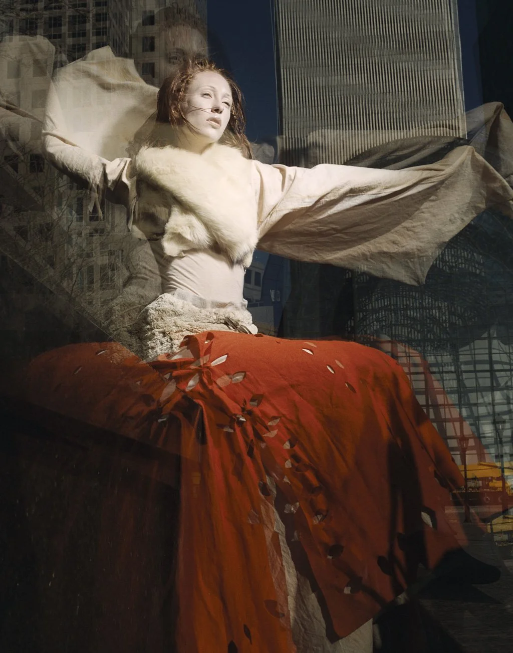 A woman lying in a display window, with her eyes closed and one arm behind her head. She is dressed in a beige outfit with a fur collar and an orange skirt, with a cityscape reflected in the glass behind her.