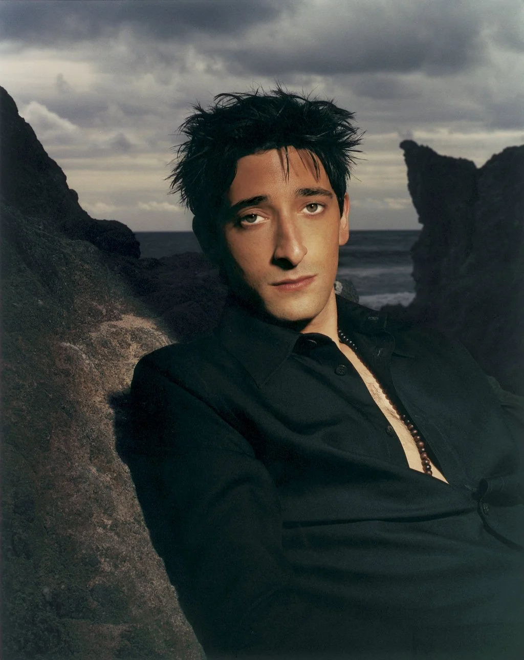 ADRIEN BRODY with spiked black hair and fair skin, gazing at the camera, wearing a black shirt partially unbuttoned, against a rocky coastline with ocean and cloudy sky in the background.