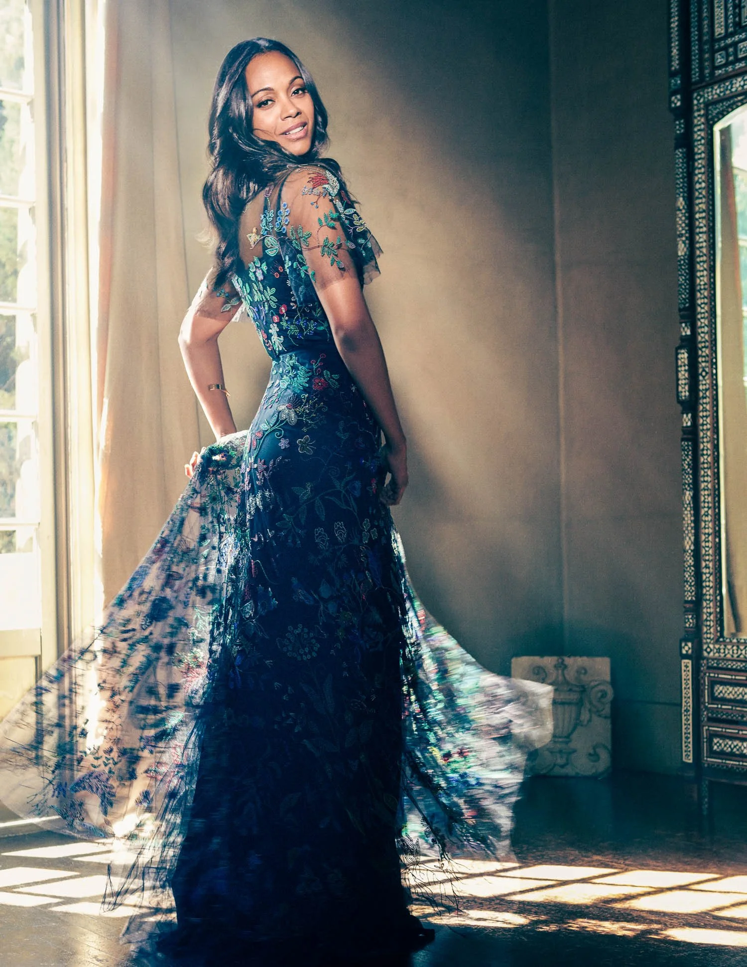 ZOE SALDANA in a sheer black dress with floral embroidery, standing near a window with sunlight coming through.