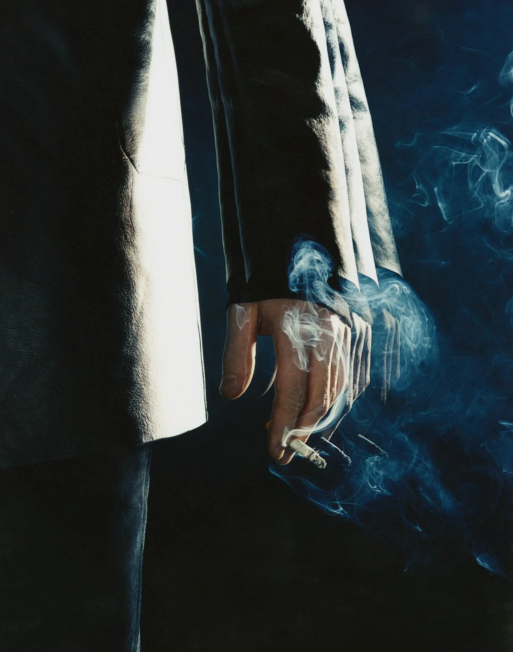 Close-up of a person wearing a black coat, holding a lit cigarette with smoke swirling around, against a dark background.