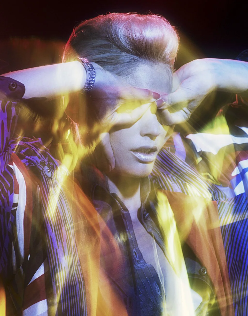 A person with styled hair, wearing a striped shirt and a bracelet, poses with arms raised and hands covering eyes, amidst colorful light effects.
