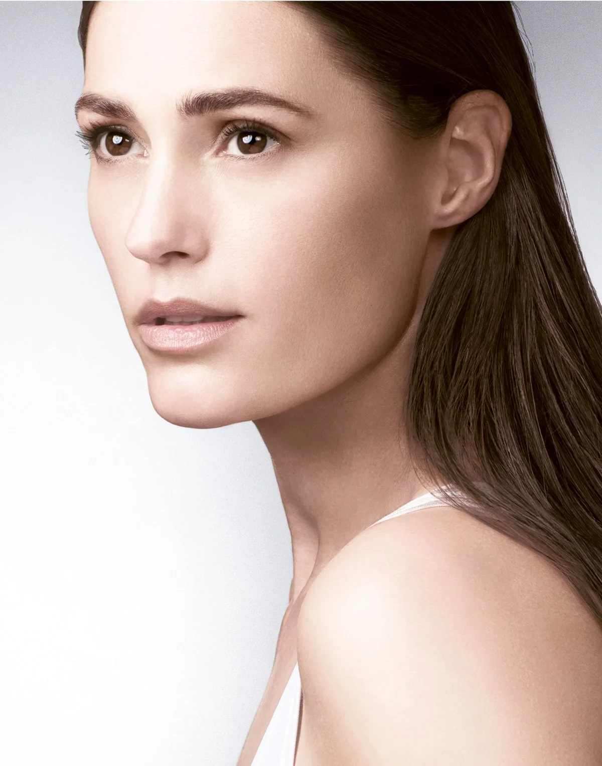 Close-up portrait of a woman with clear skin, brown hair, brown eyes, and neutral makeup, looking slightly to the side against a white background.