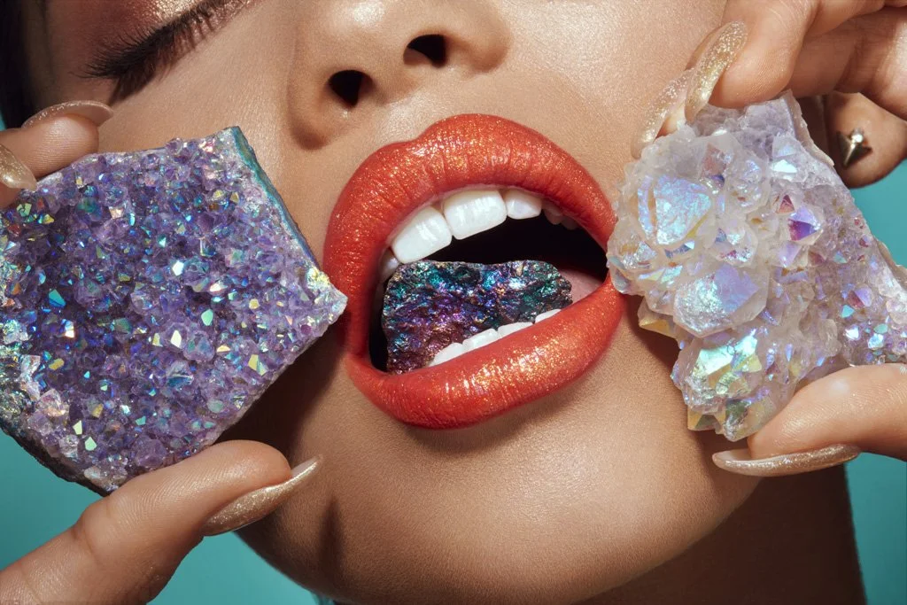 Close-up of a woman with bright orange lipstick, white teeth, holding two large iridescent mineral rocks, with a colorful rock on her tongue.