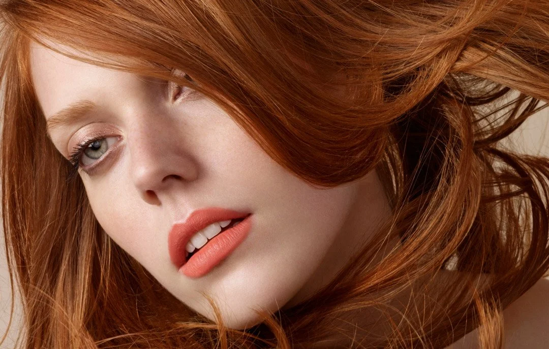 Close-up of a woman with long red hair, fair skin, and natural makeup, looking to the side with a soft expression.