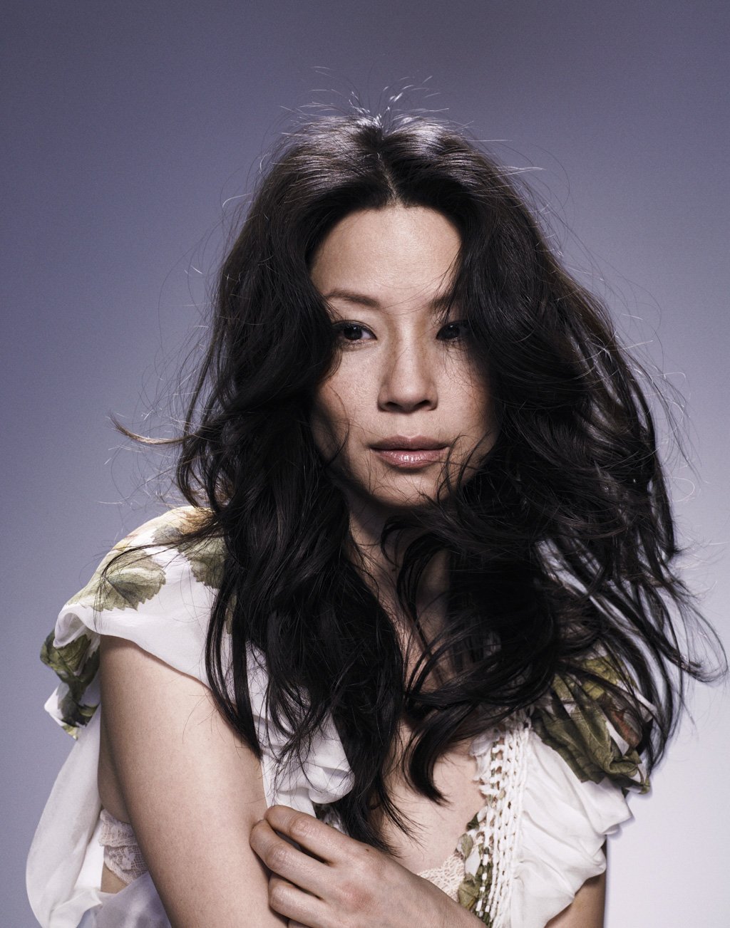 LUCY LIU with long, curly black hair and light skin looks into the camera with a neutral expression. She is wearing a white dress with floral green accents .