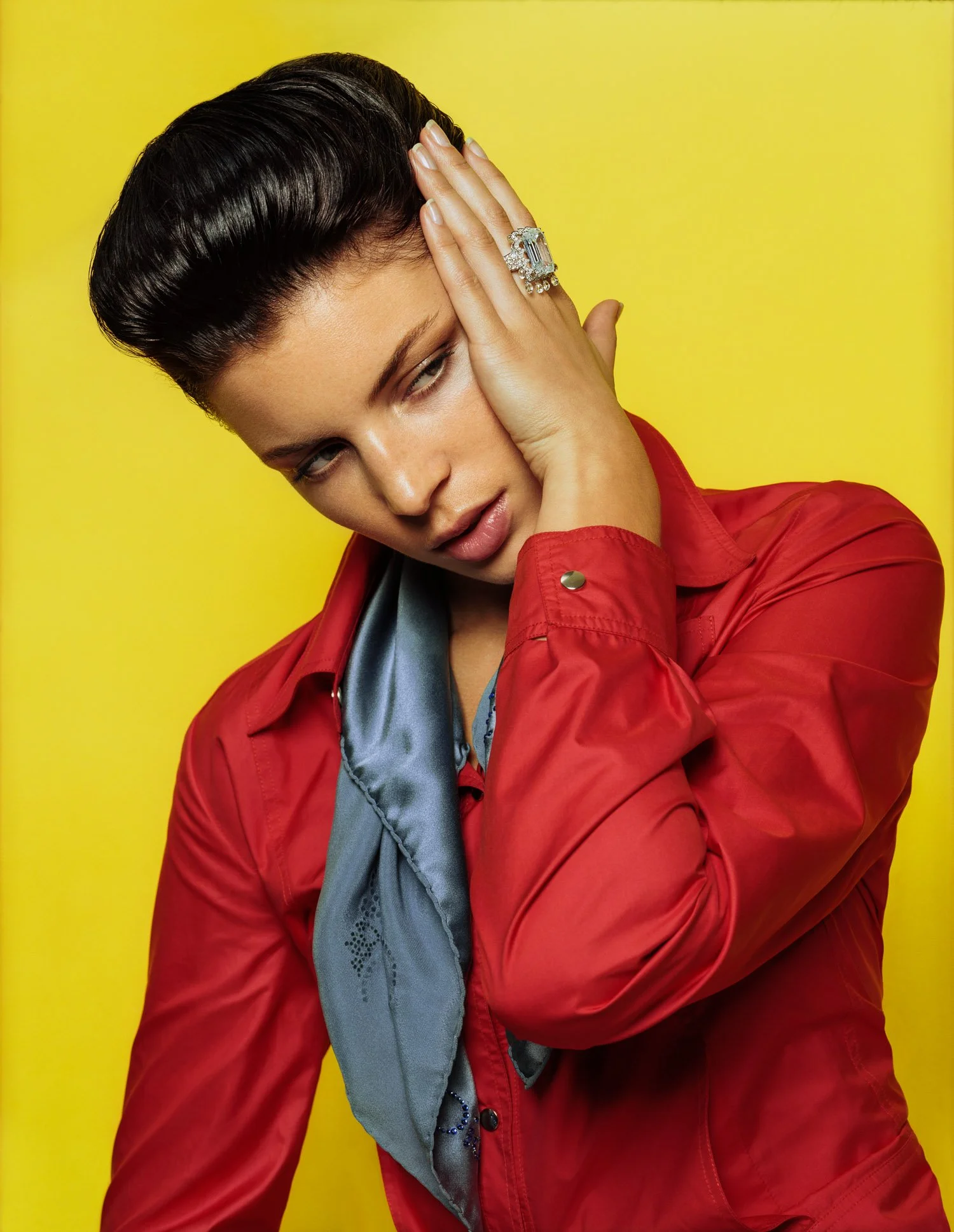 A woman with styled dark hair wearing a red jacket, a gray silk scarf, and a large statement ring, posing with her hand on her face against a yellow background.