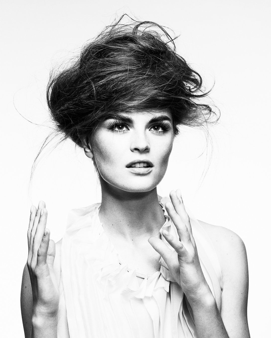 Black and white photograph of a woman with voluminous, messy hair and intense gaze, wearing a light-colored blouse with ruffles, looking directly at the camera.