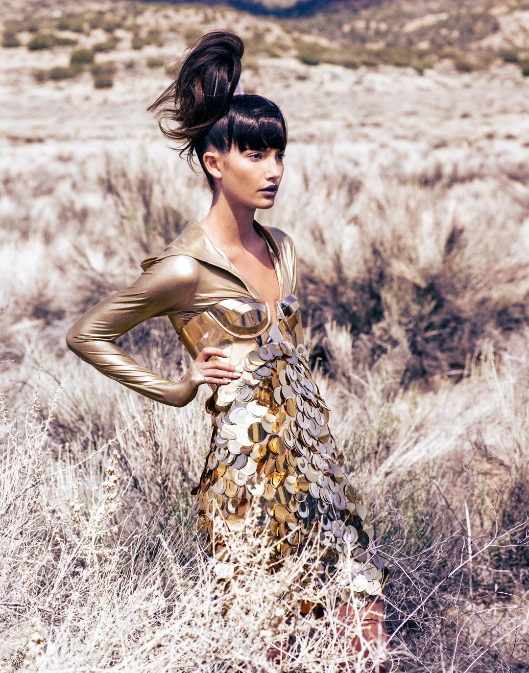 A woman standing in a dry, desert landscape with beige shrubs, wearing a shiny gold dress with large circular metallic embellishments.