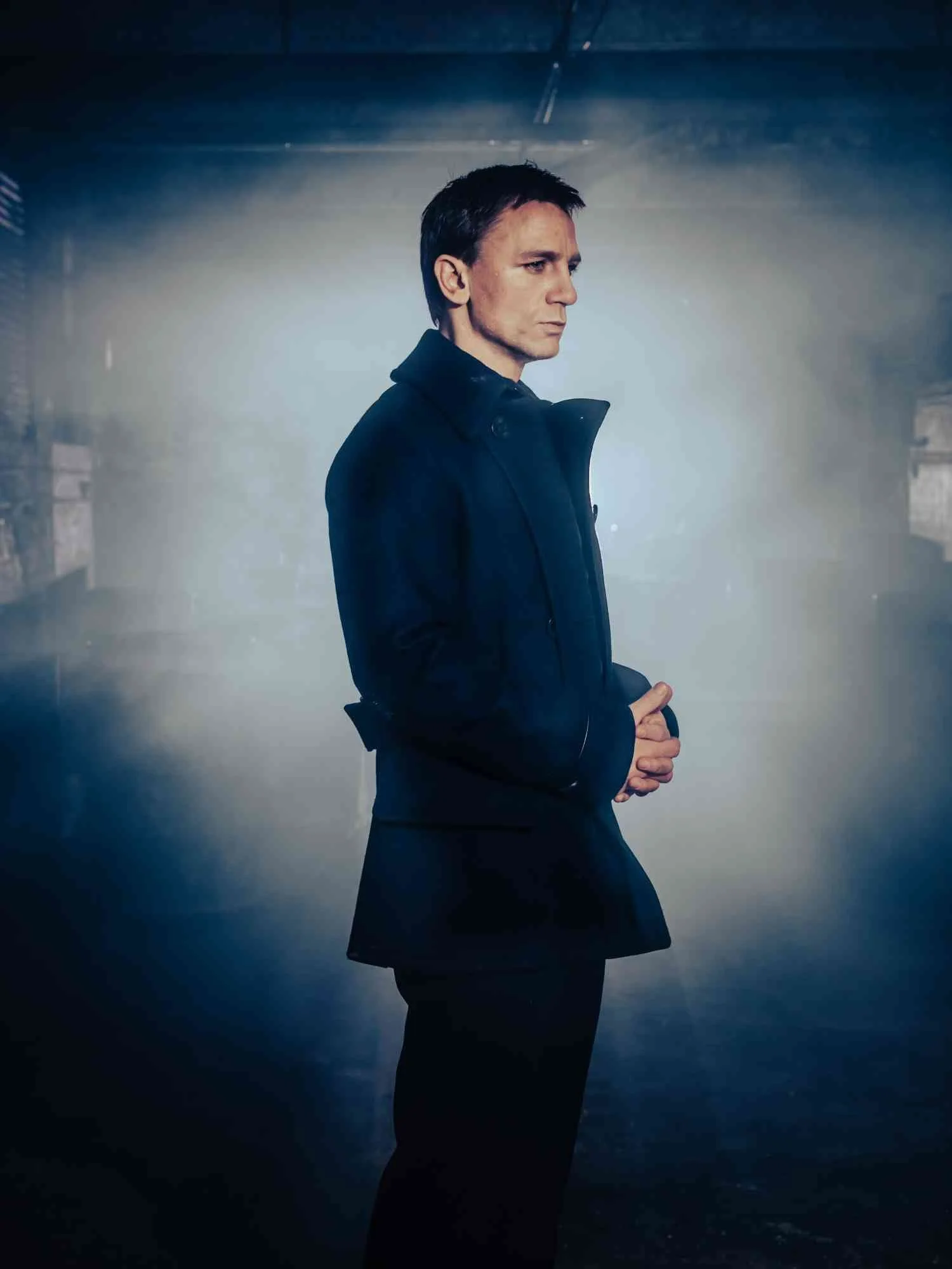 DANIEL CRAIG in a black coat stands in profile against a dim, foggy background, with his hands clasped in front of him and a serious expression on his face.