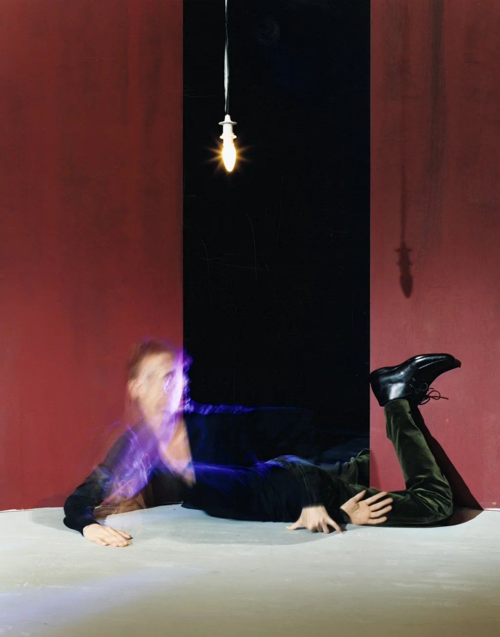 A person is lying on the floor with their upper body propped up on one arm, blurred motion suggests movement or a quick gesture. The shadow of a hanging light bulb is cast on a red wall behind them, with a black opening in the wall. The person is wea