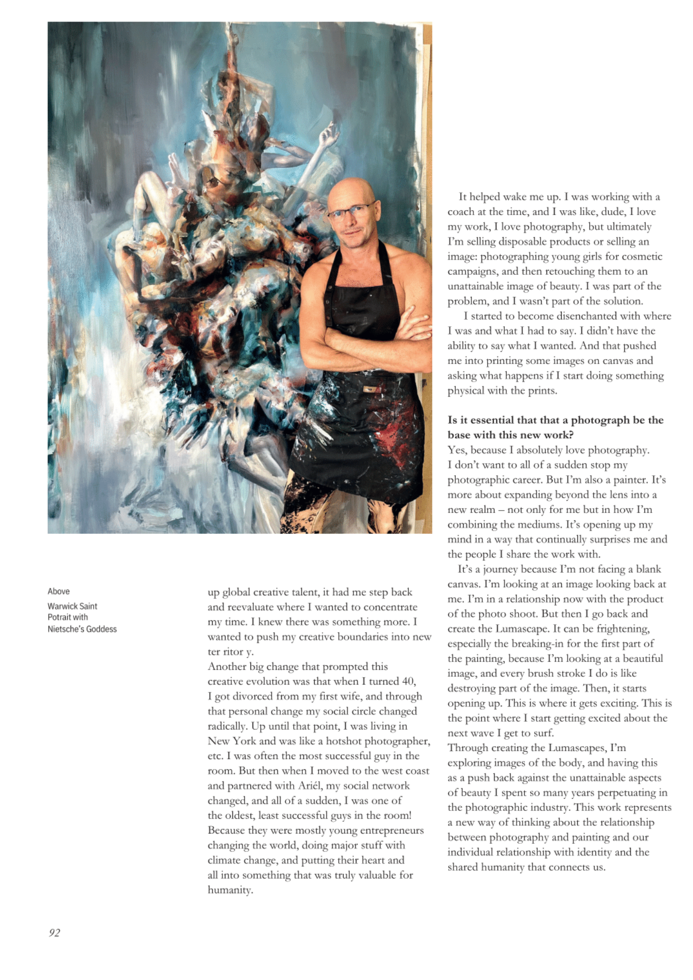 A man in a black apron with paint stains stands confidently with his arms crossed in front of a large, expressive abstract painting of a goddess or mythological figure with multiple arms on a textured blue background.