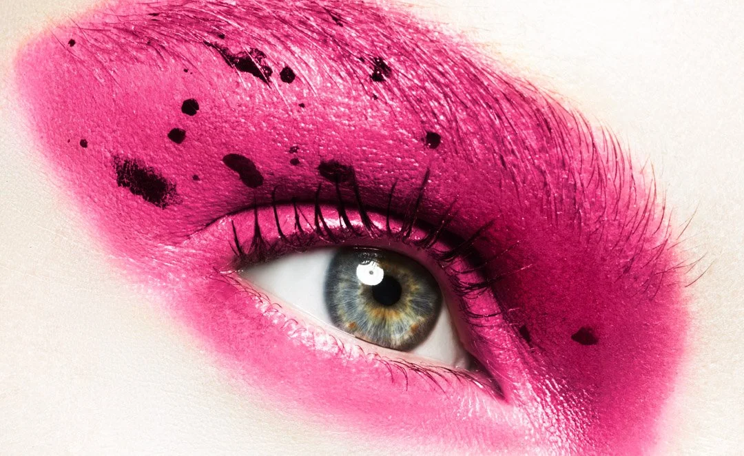 Close-up of a human eye with vibrant pink eye makeup and black ink splatters on the eyelid.