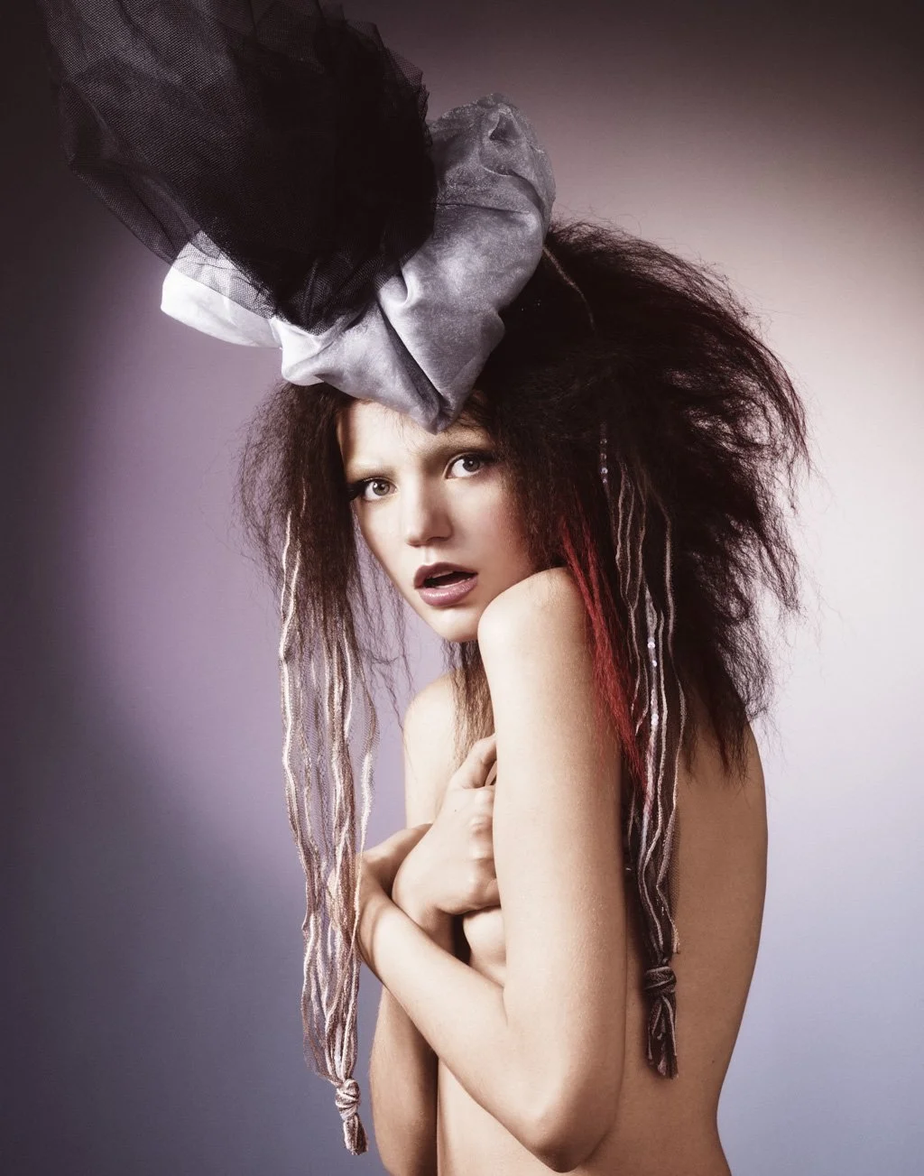 A topless woman with messy, voluminous hair with red streaks, holding her arms across her chest, wearing a large decorative hat with black and gray netting, against a gradient background.