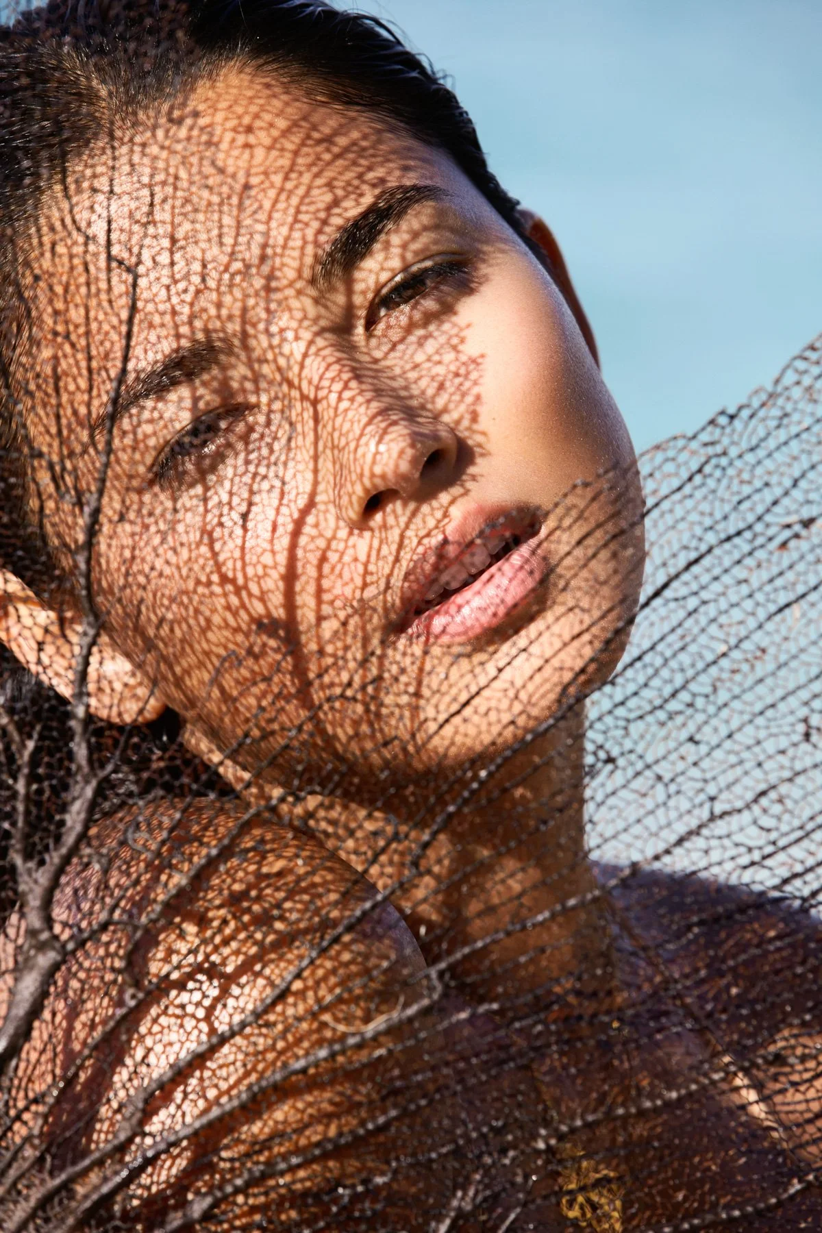 Close-up of a woman's face with her eyes partially closed, behind a delicate, intricate leaf skeleton casting shadows on her face, against a blue sky background.