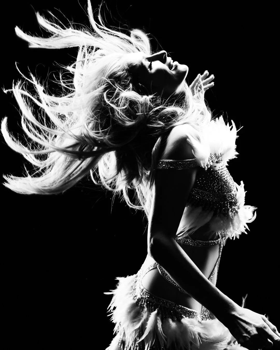 A black and white image of PARIS HILTON dancing with her hair flowing, wearing a feathered and rhinestone-studded outfit.
