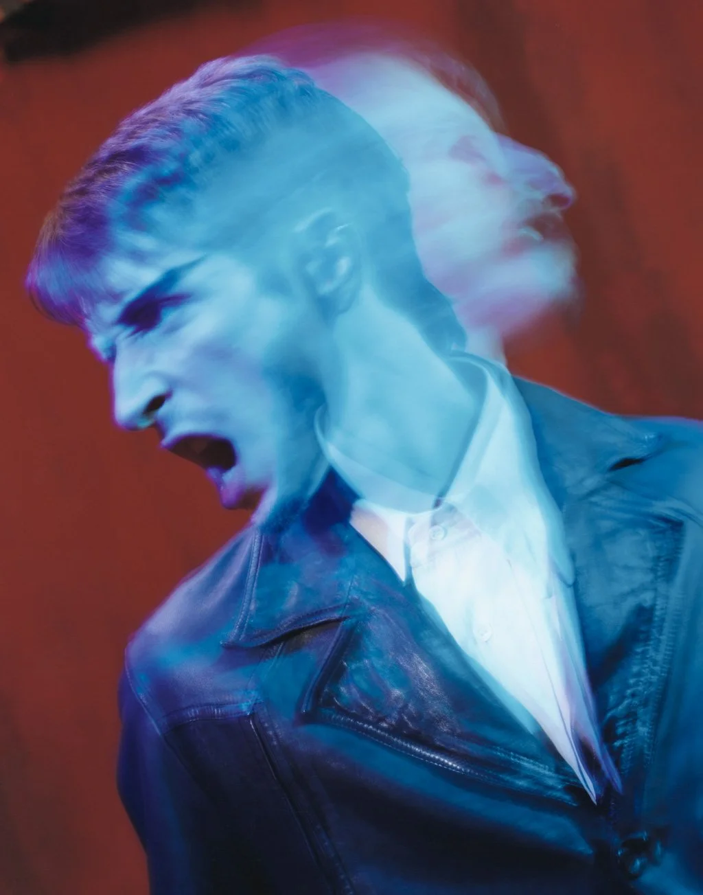 A man with short hair and a beard wearing a leather jacket and a white shirt, with a motion blur effect giving a double exposure look against a red background.
