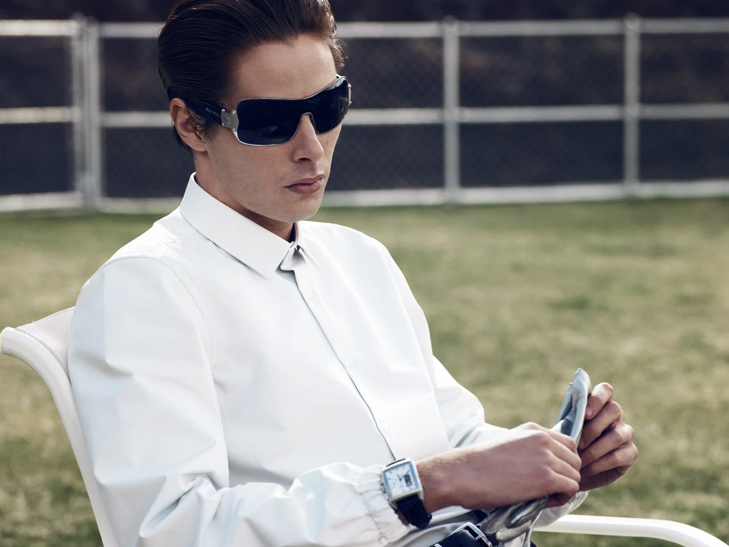 Person with short dark hair wearing large dark sunglasses, a white shirt, and a smartwatch, sitting outdoors and holding a smartphone.